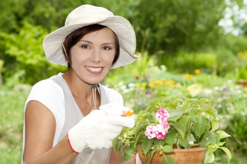 A woman wearing a wide-brimmed white gardening hat, white gloves, and a light grey top is smiling while tending to a potted flowering plant with pink blossoms, set in a lush garden area. The garden features a well-maintained lawn with dense green grass in the foreground, and a variety of blurred shrubs, flower beds, and trees in the background, indicating a vibrant outdoor space typical of a residential garden in Brixton. The scene is evenly lit, suggesting a bright, possibly partly cloudy day, with natural sunlight illuminating the greenery and flowers. This setting emphasizes outdoor gardening activities and landscape care, consistent with professional garden maintenance services provided by companies like Garden Maintenance Brixton, supporting natural landscape aesthetics in London suburbs.