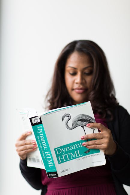 A woman with dark brown hair, dressed in a black blazer over a burgundy top, is holding and reading a book titled 'Dynamic HTML' by O'Reilly, which features a realistic illustration of a flamingo on t